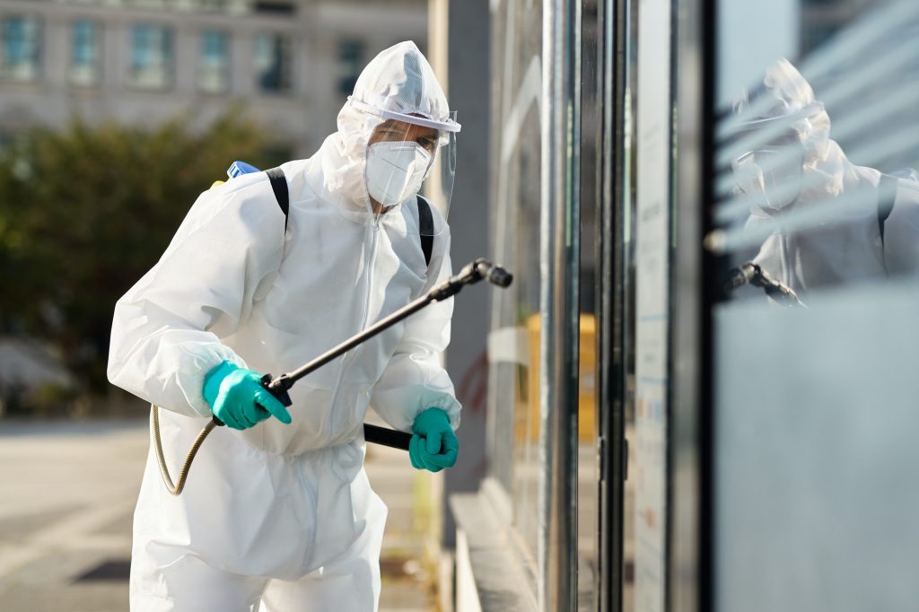 Sanitation worker in hazmat suit disinfecting public building during coronavirus epidemic.