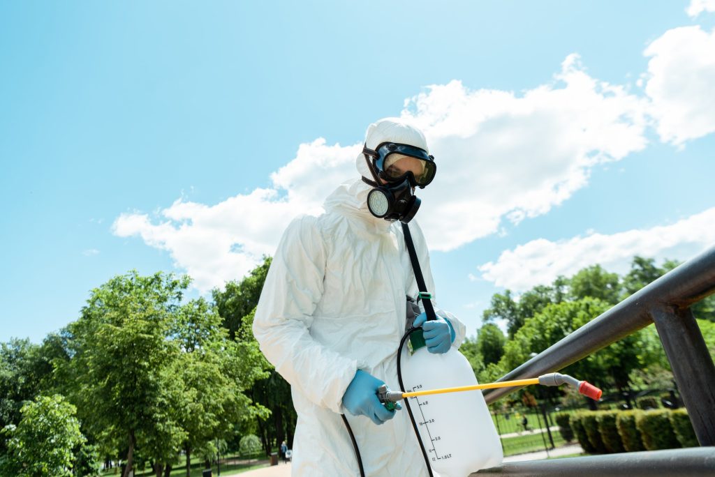 specialist in hazmat suit and respirator disinfecting railings in park during covid-19 pandemic